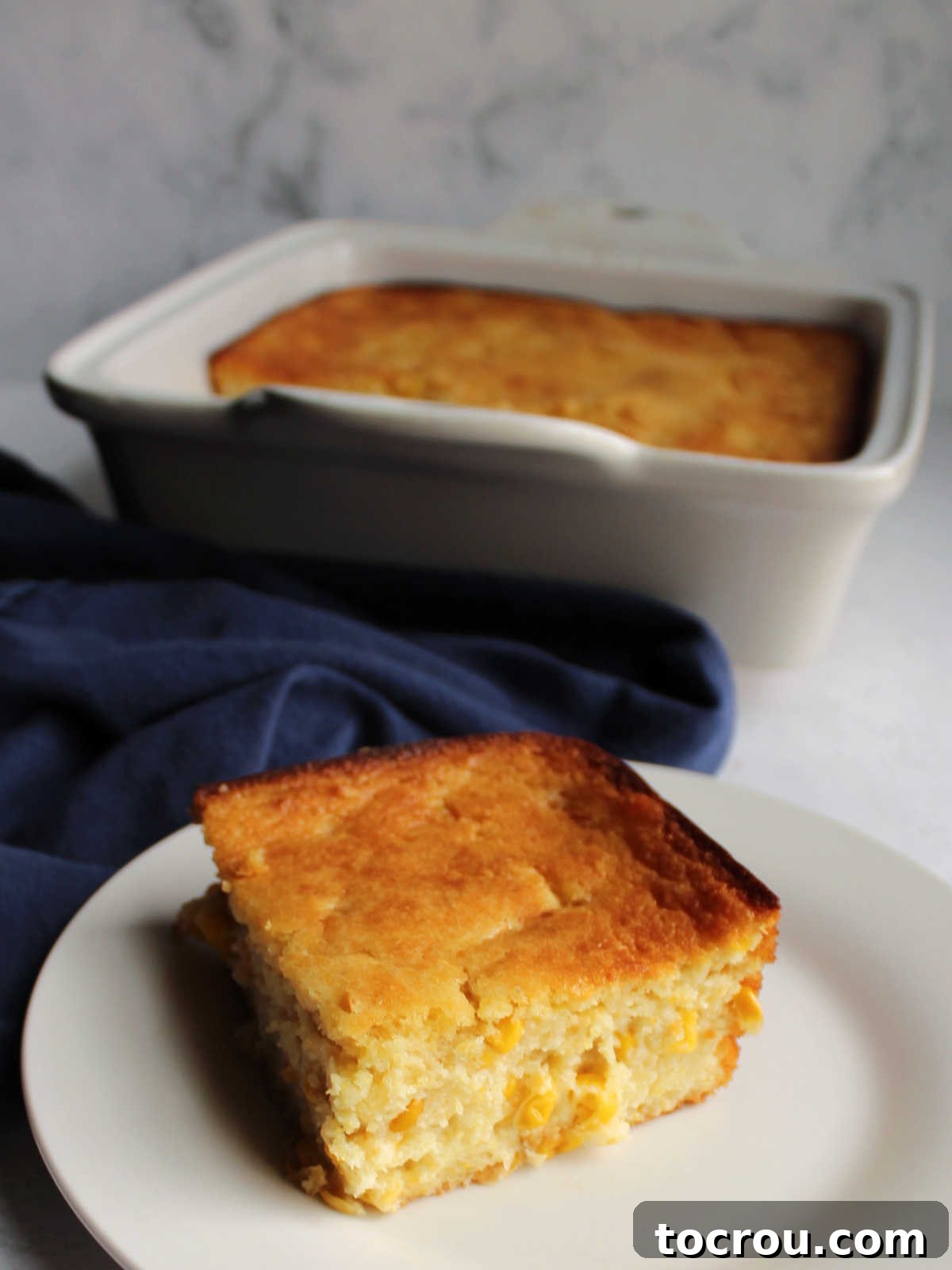 Golden Corn Delight 5 A piece of corn casserole on a plate, with the remaining corn pudding in a square baking dish in the background.