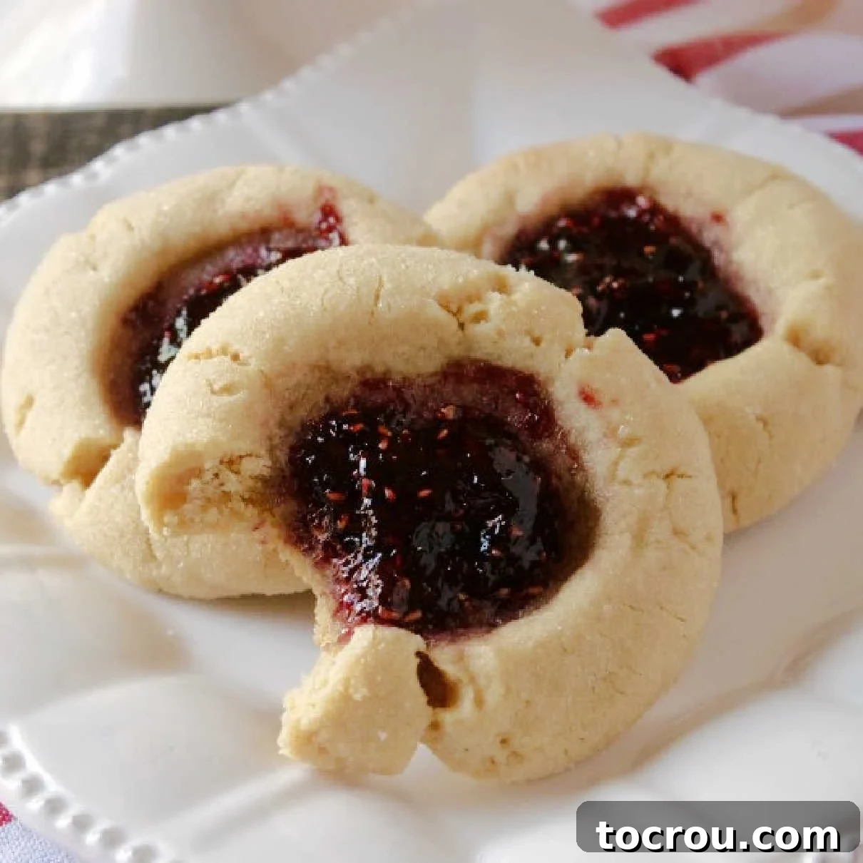 Jelly-Kissed Peanut Butter Bites 2 Plate with three peanut butter and jelly thumbprint cookies, one with a bite taken out, showcasing the jam filling.
