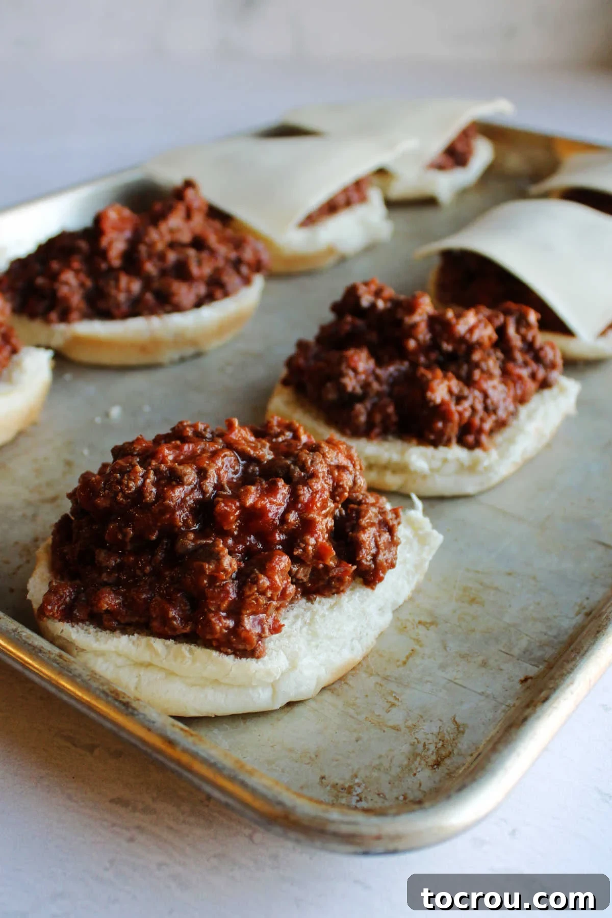 Hamburger buns lined on a baking tray, generously topped with the pizza burger meat mixture and mozzarella cheese, ready for baking.