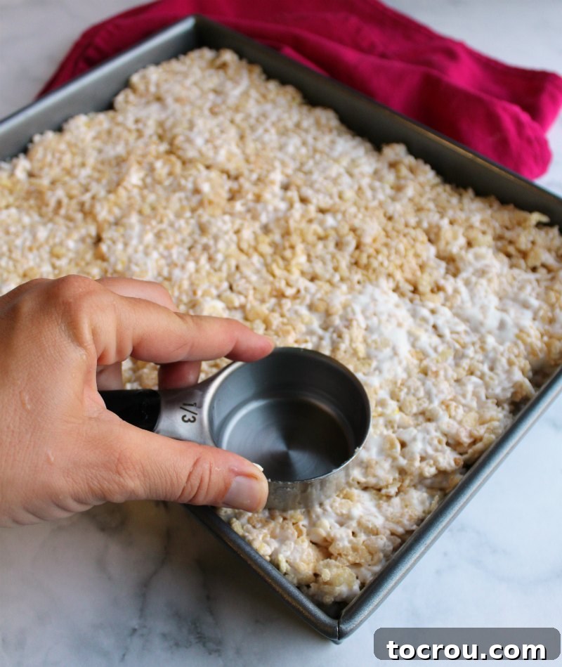 Pressing rice krispie treats down firmly into a baking pan using a greased measuring cup to ensure a compact, even layer.