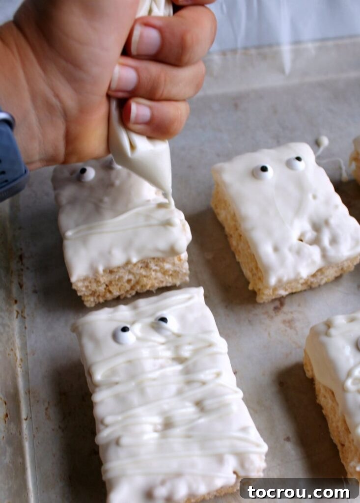 A hand piping melted white chocolate in jagged lines onto rice crispy treats, creating the bandage effect for mummies.