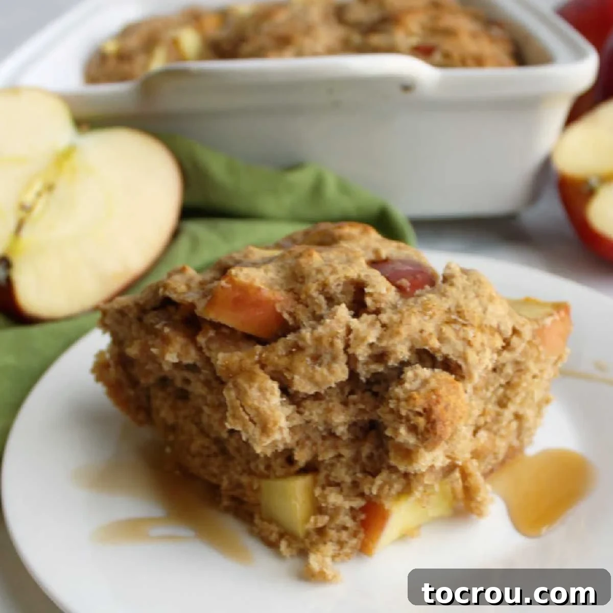 Close-up slice of rustic buttermilk apple cake, featuring juicy apple chunks, with a warm, inviting backdrop of apple halves and a cake pan.