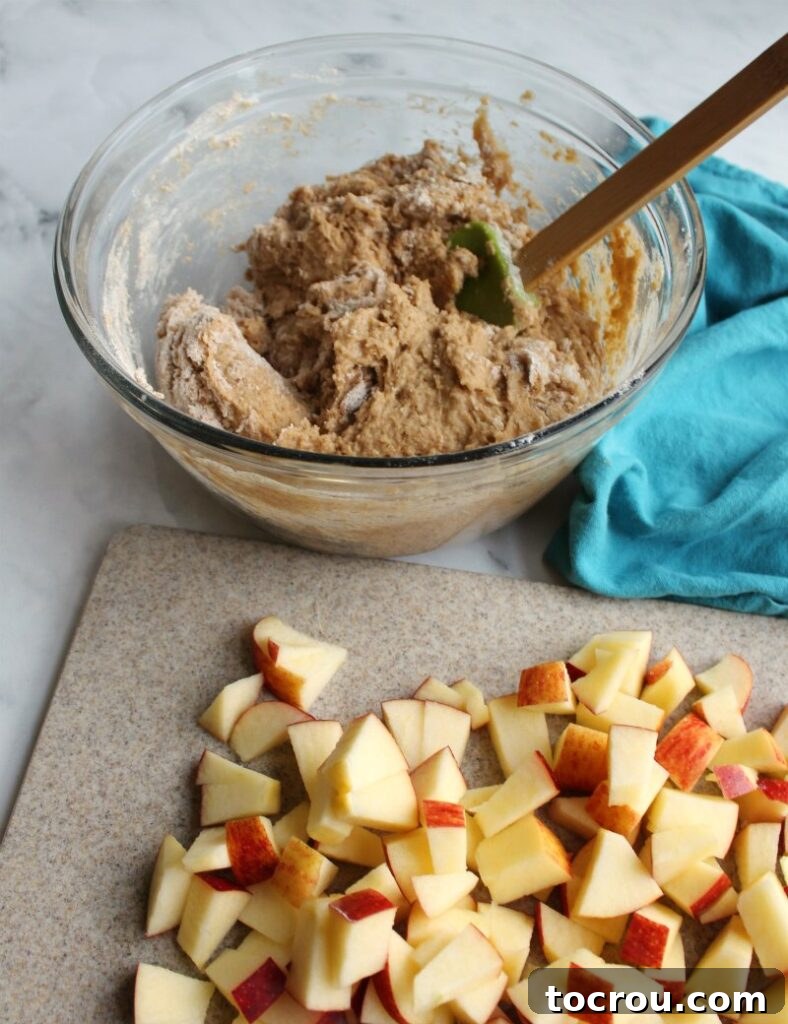 A bowl filled with luscious apple cake batter positioned behind a cutting board generously covered with freshly chopped apples, ready to be incorporated.