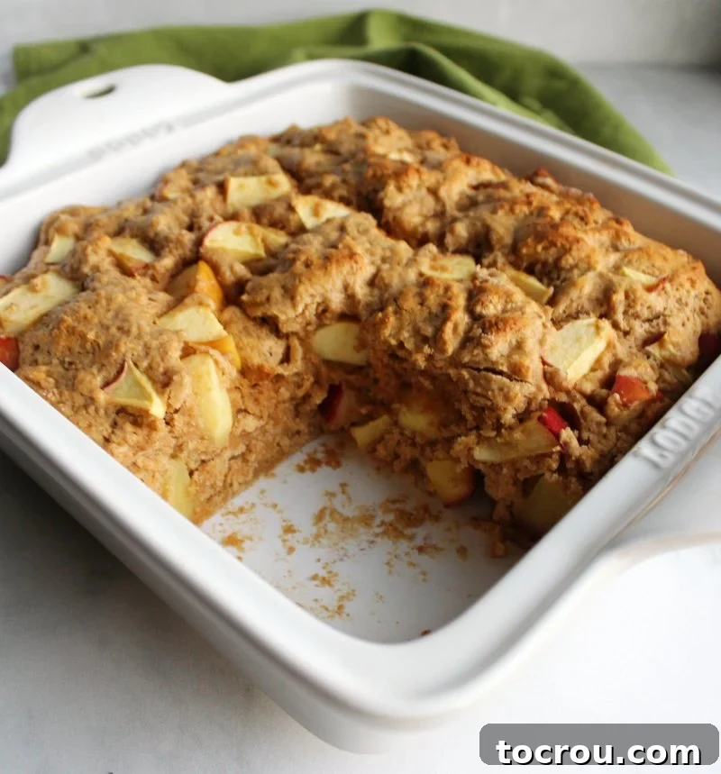 A square pan of rustic buttermilk apple cake with a perfectly cut corner piece missing, showcasing its moist texture and apple chunks.