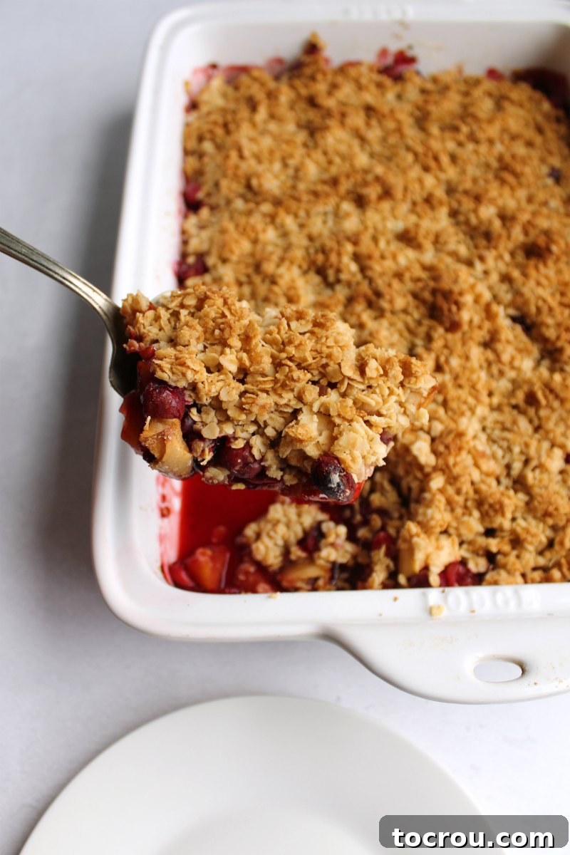Serving spoon filled with cranberry apple crisp with golden oat topping being lifted out of the baking pan.