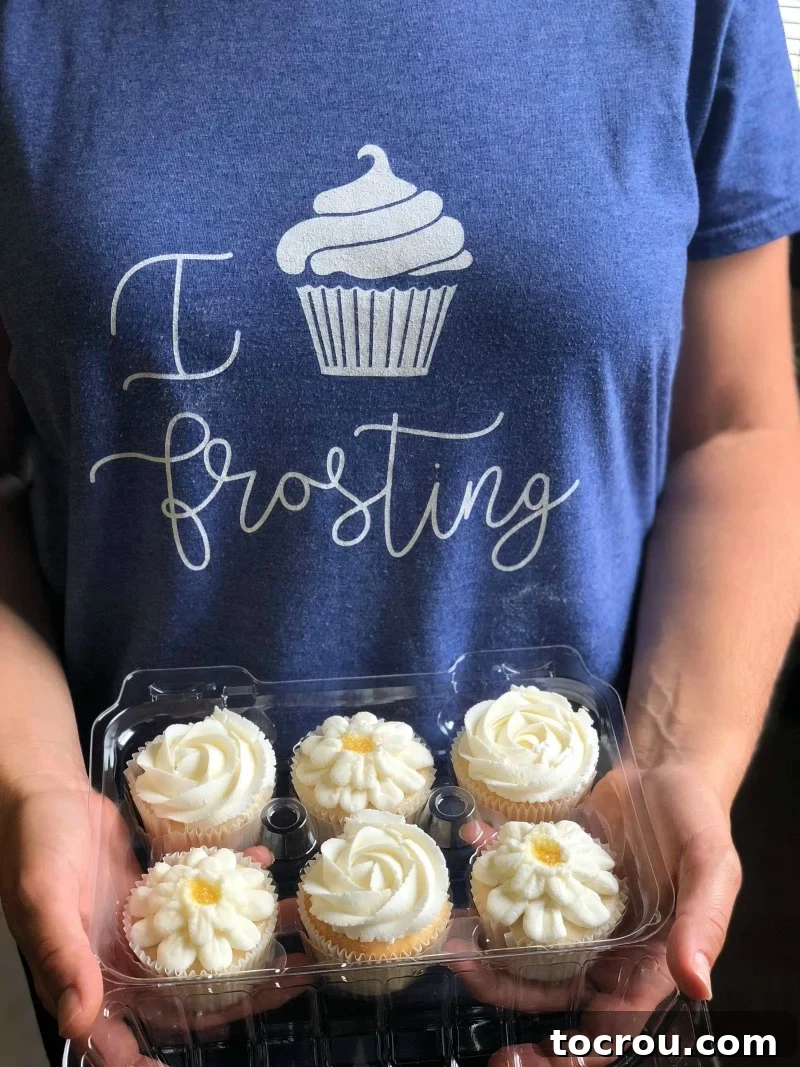 A pair of hands delicately holding a clear container filled with beautifully decorated cupcakes, each topped with intricate buttercream flower designs in various pastel shades.