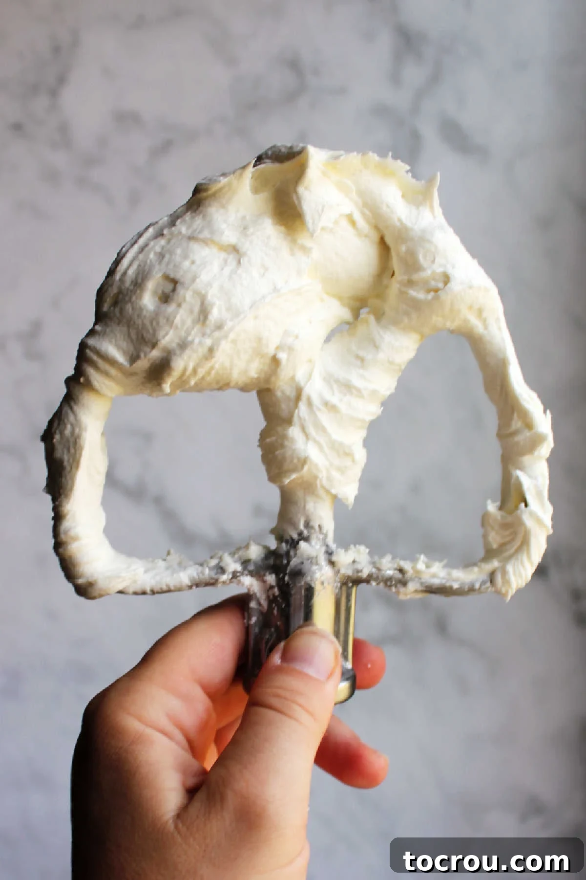 A hand holding a mixer paddle laden with a generous scoop of fluffy, pristine white buttercream frosting, showcasing its smooth and airy texture.