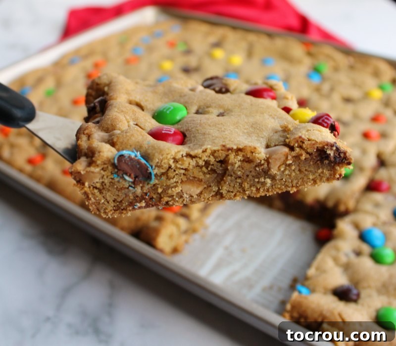 Chewy brown sugary blondies loaded with chocolate chunks, caramel undertones and topped with M&Ms. This big batch of bars is perfect for a party, carry-in or just because. first piece salted caramel M&M blondies lifted out of pan on spatula showing chewy fudgy center.