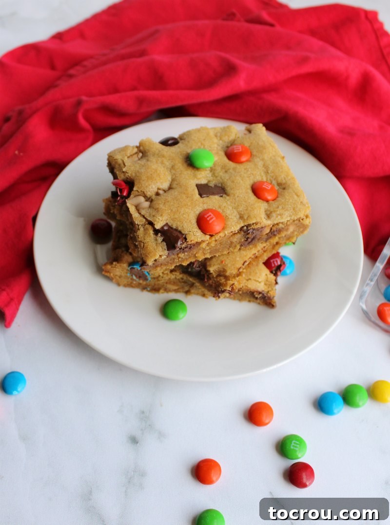 Chewy brown sugary blondies loaded with chocolate chunks, caramel undertones and topped with M&Ms. This big batch of bars is perfect for a party, carry-in or just because. looking down on a couple of pieces of chewy salted caramel m&m blondies on plate with candies scattered around.