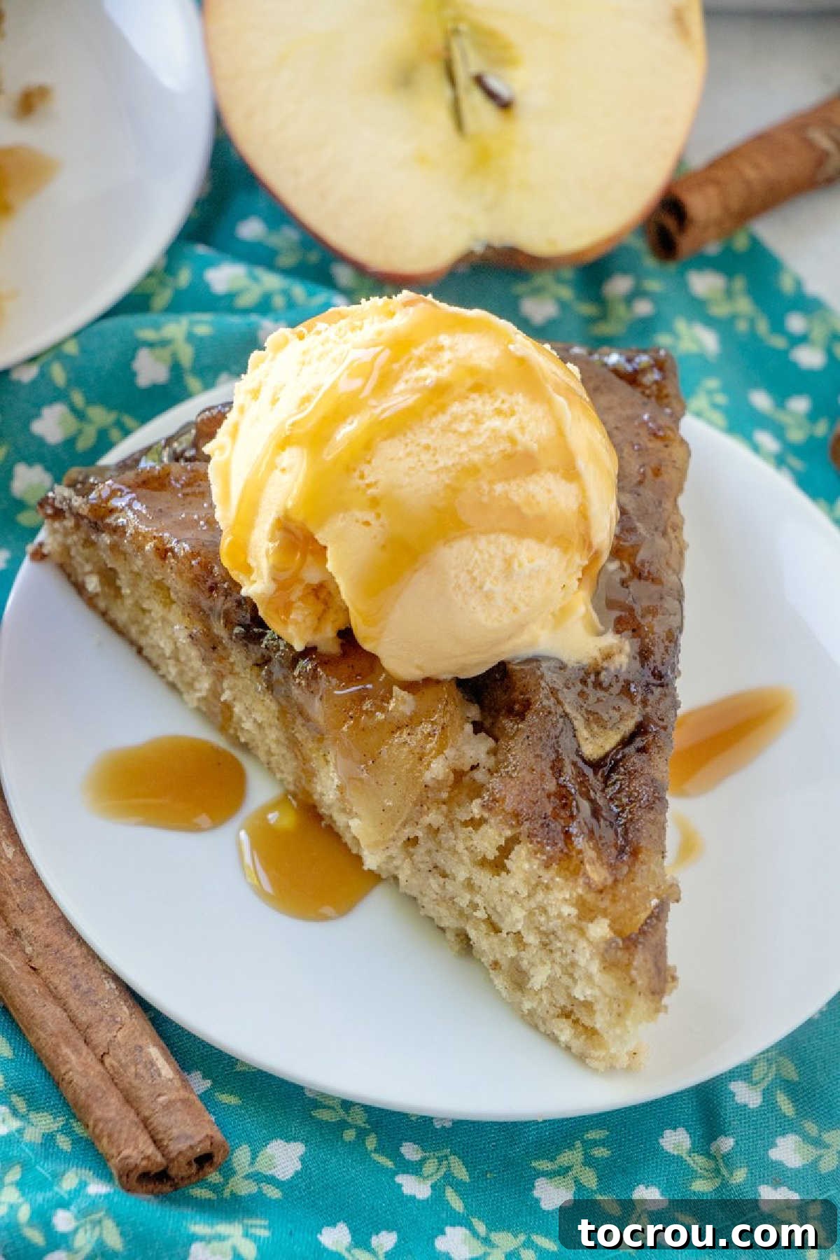 Gooey Caramel Apple Upside Down Cake 3 Looking down on slice of caramel apple upside down cake topped with a scoop of vanilla ice cream and a drizzle of caramel sauce with cinnamon sticks and fresh apples in the background.