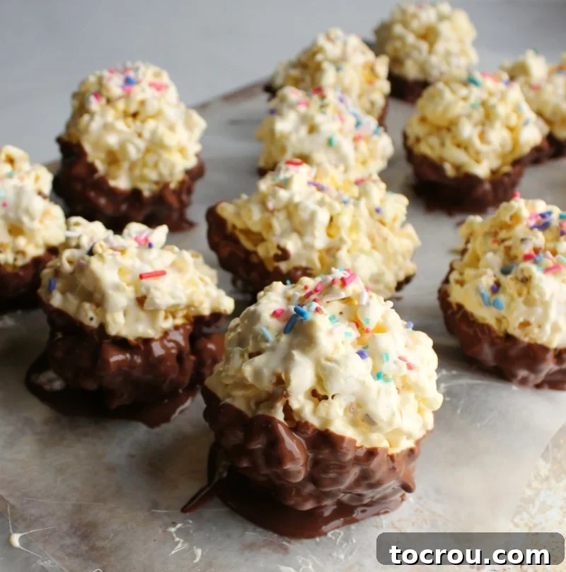 Marshmallowy Popcorn Delights 2 Marshmallow popcorn balls with the bottom halves dipped in chocolate and sprinkles on top setting up on a sheet of wax paper.