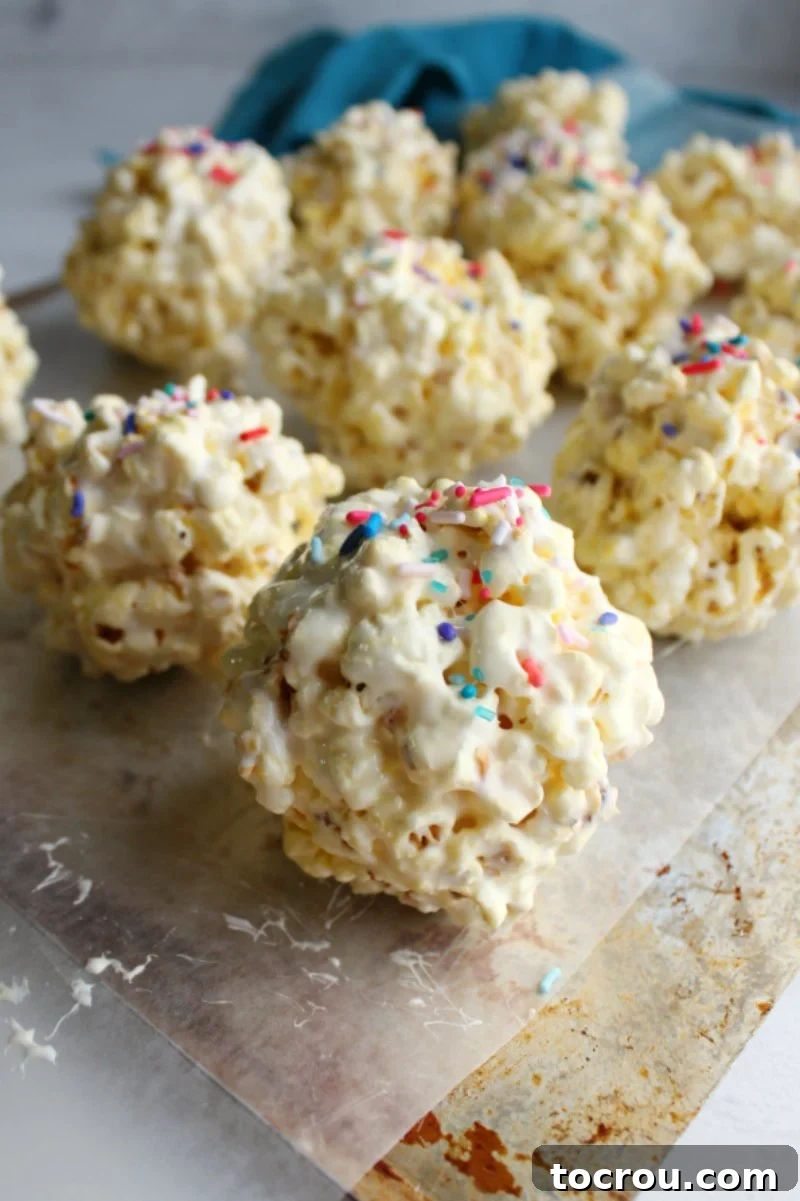 Marshmallowy Popcorn Delights 5 close up of marshmallow popcorn balls with sprinkles on top sitting on wax paper lined baking tray.