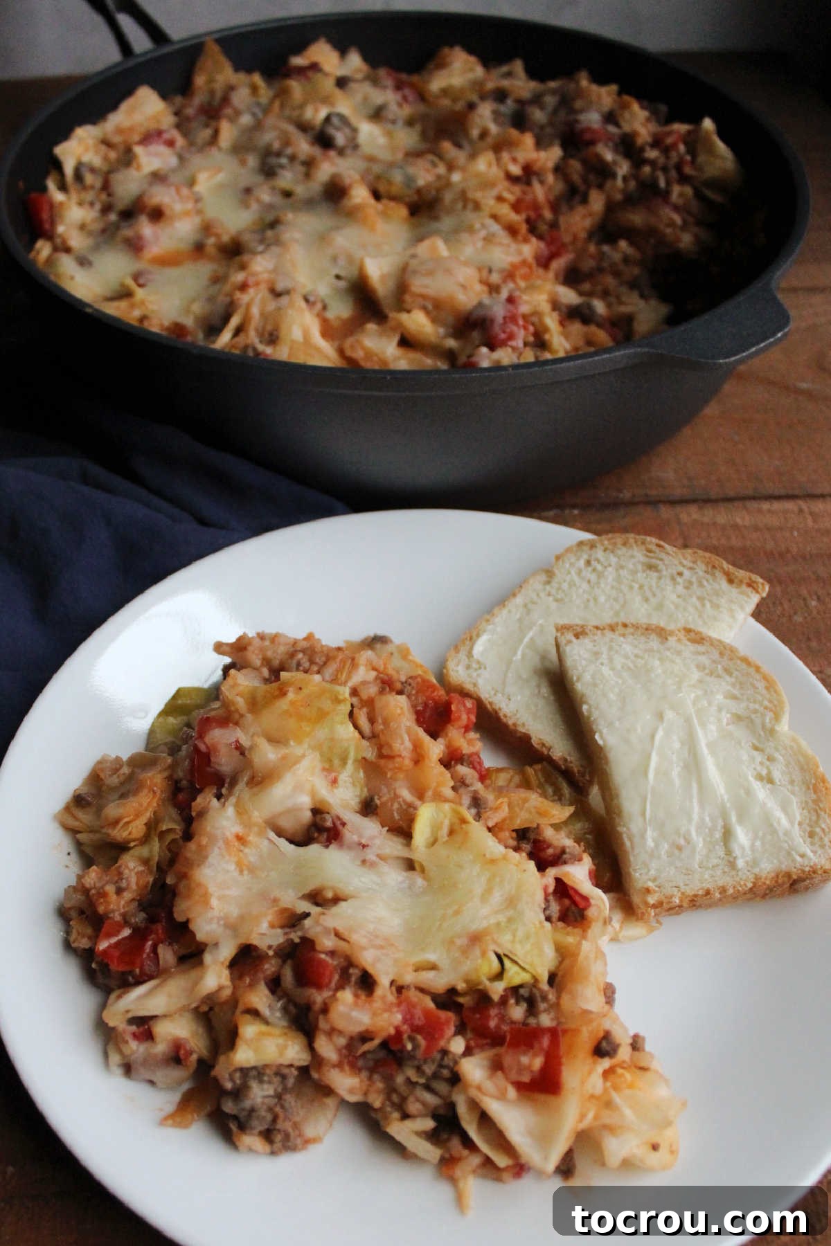 A dinner plate featuring a serving of unstuffed cabbage roll skillet dinner, beautifully topped with melted cheese and served alongside a slice of buttered bread.