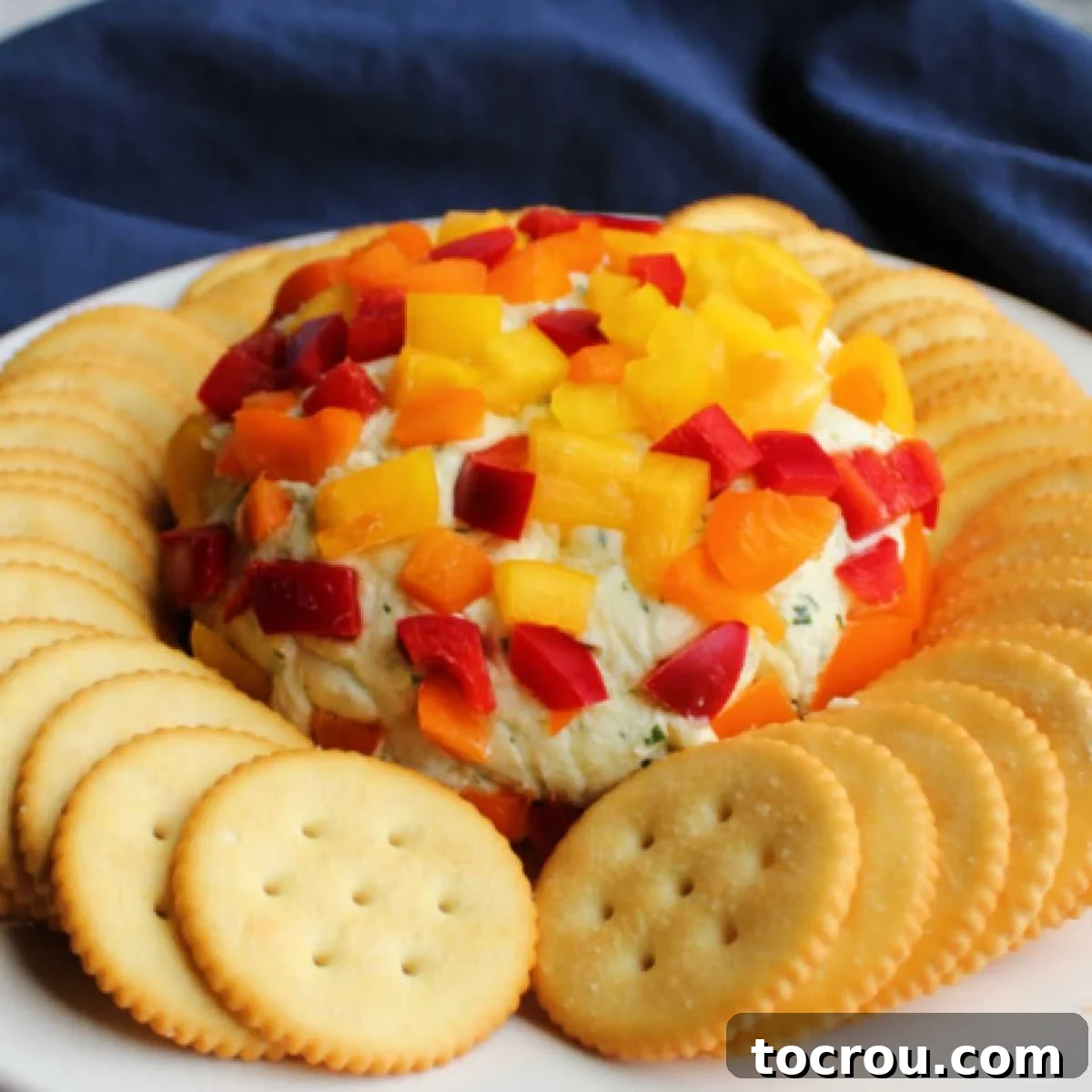 Creamy havarti cheese ball coated in diced red, yellow, and orange bell pepper on plate surrounded by crackers, ready to be served.