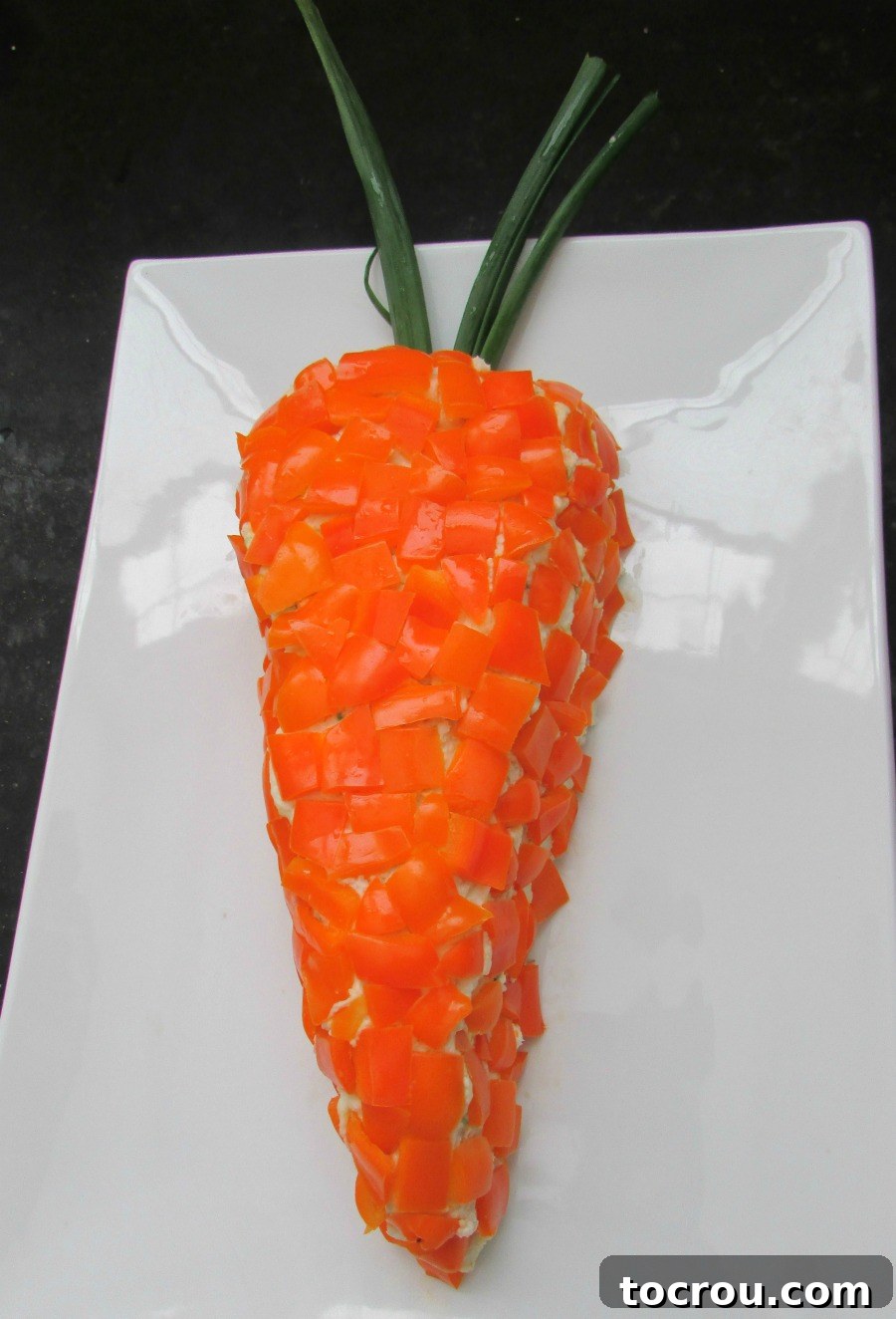 Havarti cheese ball shaped in a carrot shape with orange bell peppers on the outside and green onion greens for carrot greens.