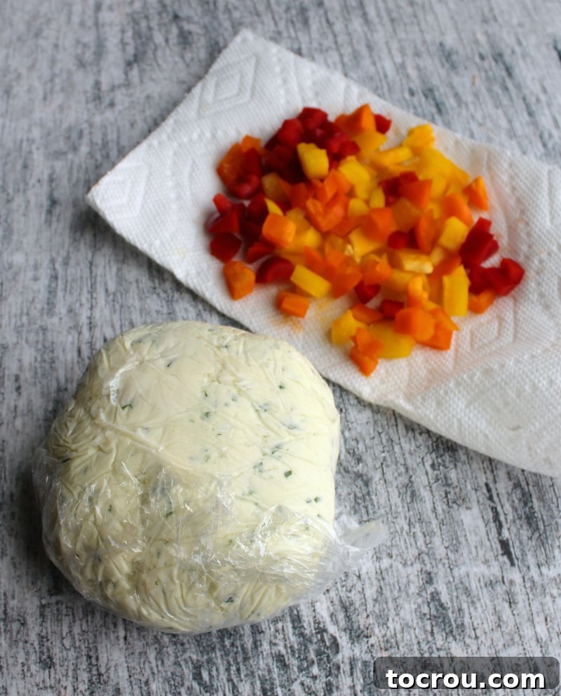 Havarti cheese ball wrapped in plastic wrap out of fridge after chilling with diced colored peppers on a paper towel behind it ready for rolling/coating.