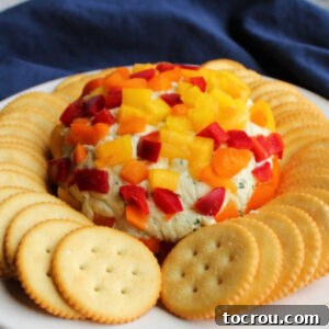 Creamy havarti cheese ball coated in diced red, yellow, and orange bell pepper on plate surrounded by crackers, ready to be served.
