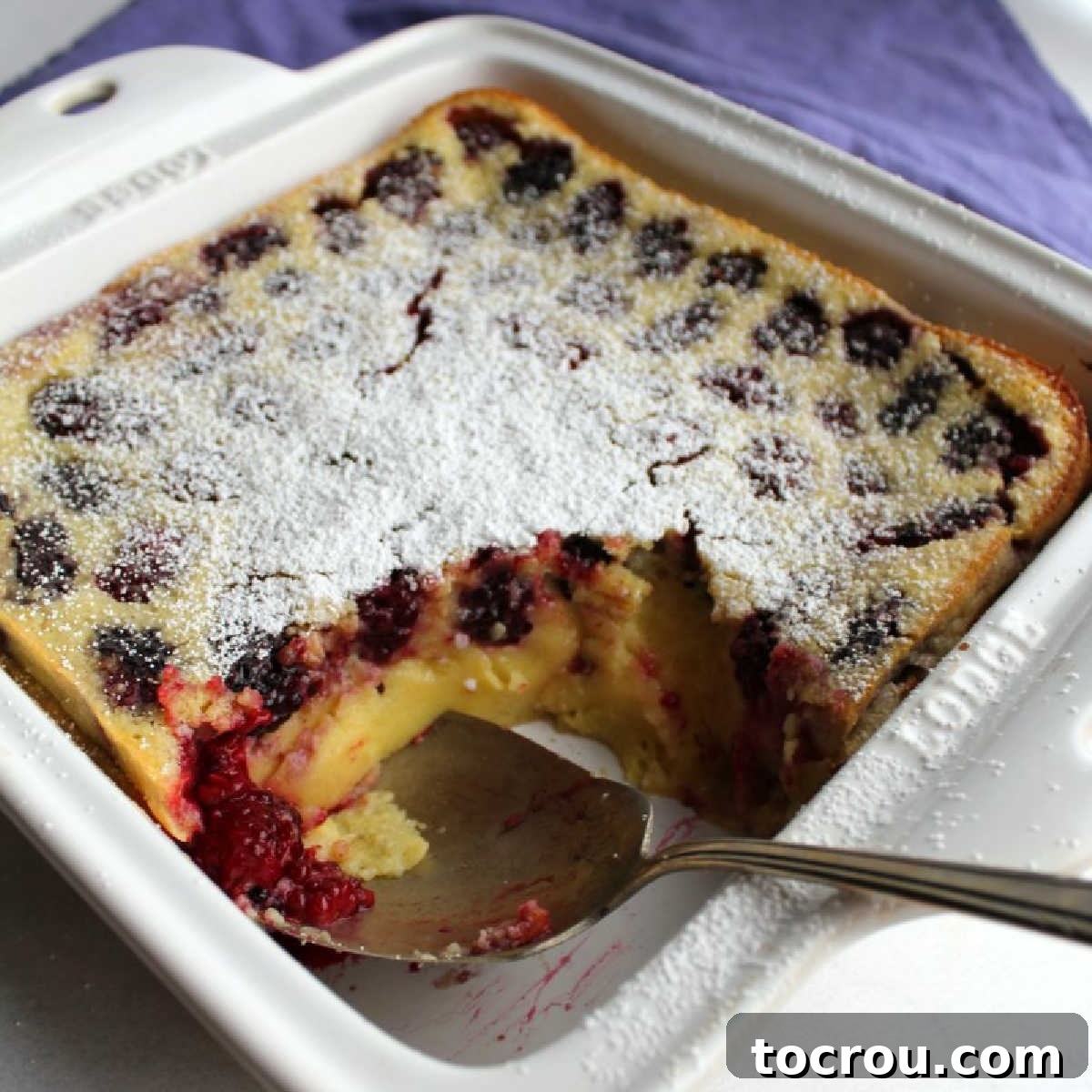 Blackberry Clafoutis with Lemon Zest Golden, custardy blackberry clafoutis in a square pan, with a berry-filled spoon hinting at the delicious inside.
