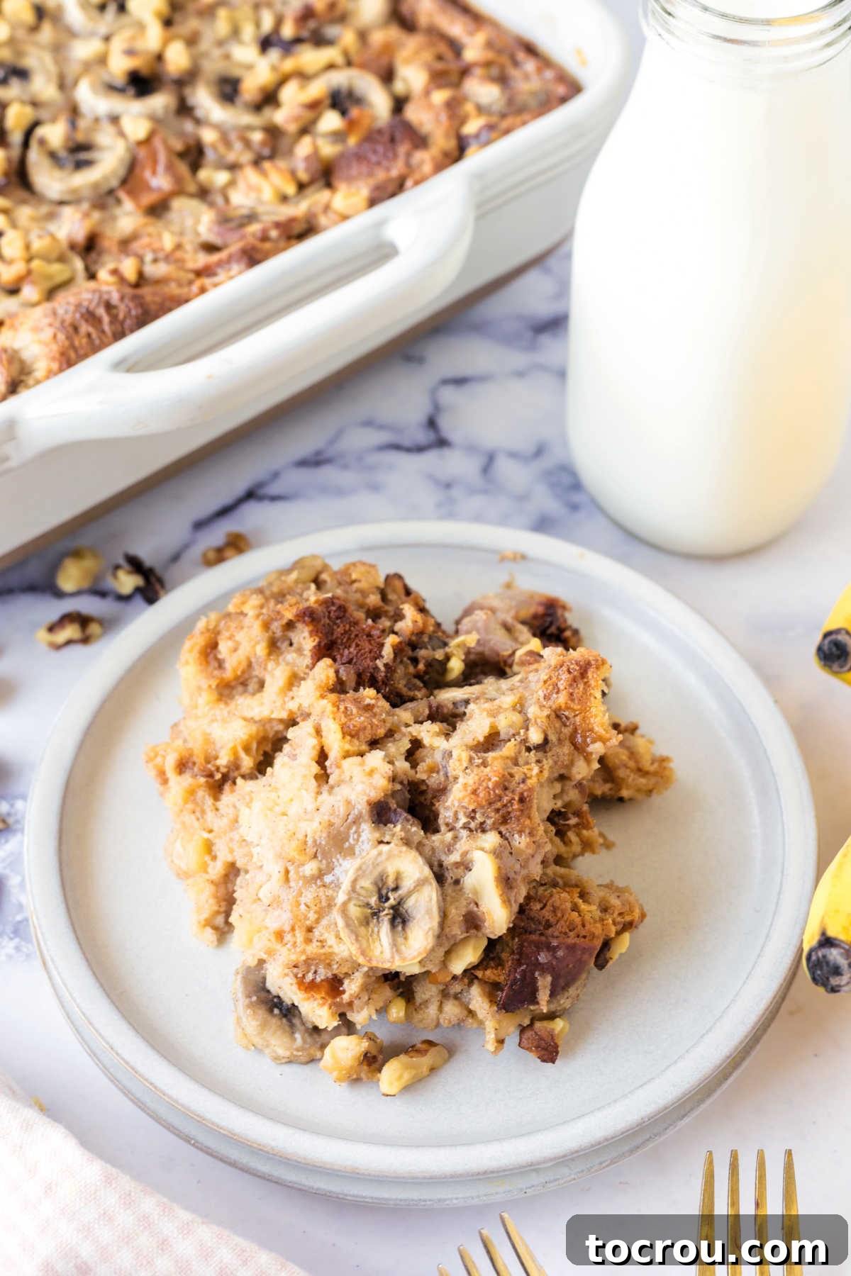 A beautifully presented serving of banana nut bread pudding on a pristine plate, with a small jug of milk and the remaining pan of pudding visible in the soft-focus background, highlighting its comforting appeal.