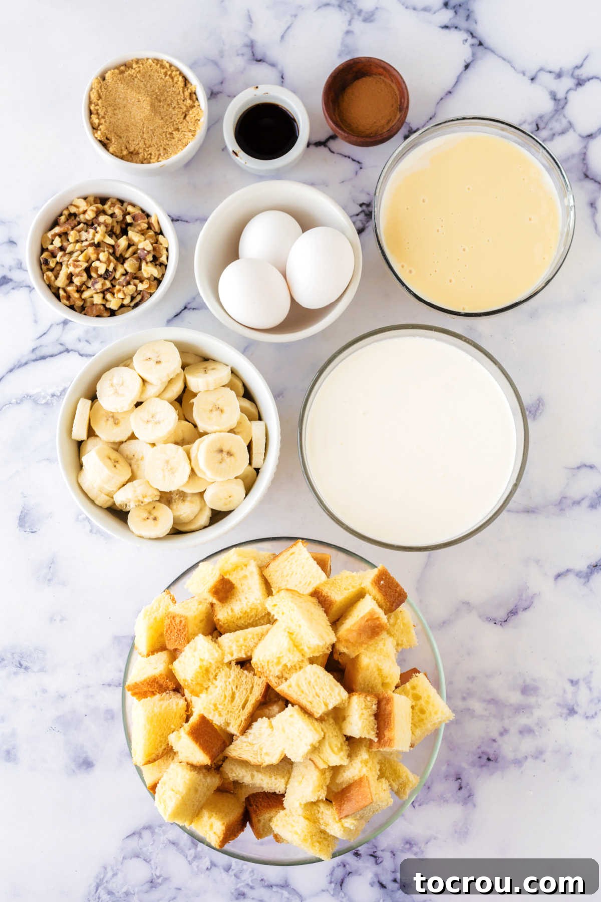 A vibrant display of fresh ingredients for banana bread pudding laid out, including perfectly cubed bread, a bottle of fresh milk, farm-fresh eggs, a can of sweetened condensed milk, artfully sliced bananas, a bowl of chopped walnuts, golden-brown sugar, a bottle of vanilla extract, and aromatic ground cinnamon.