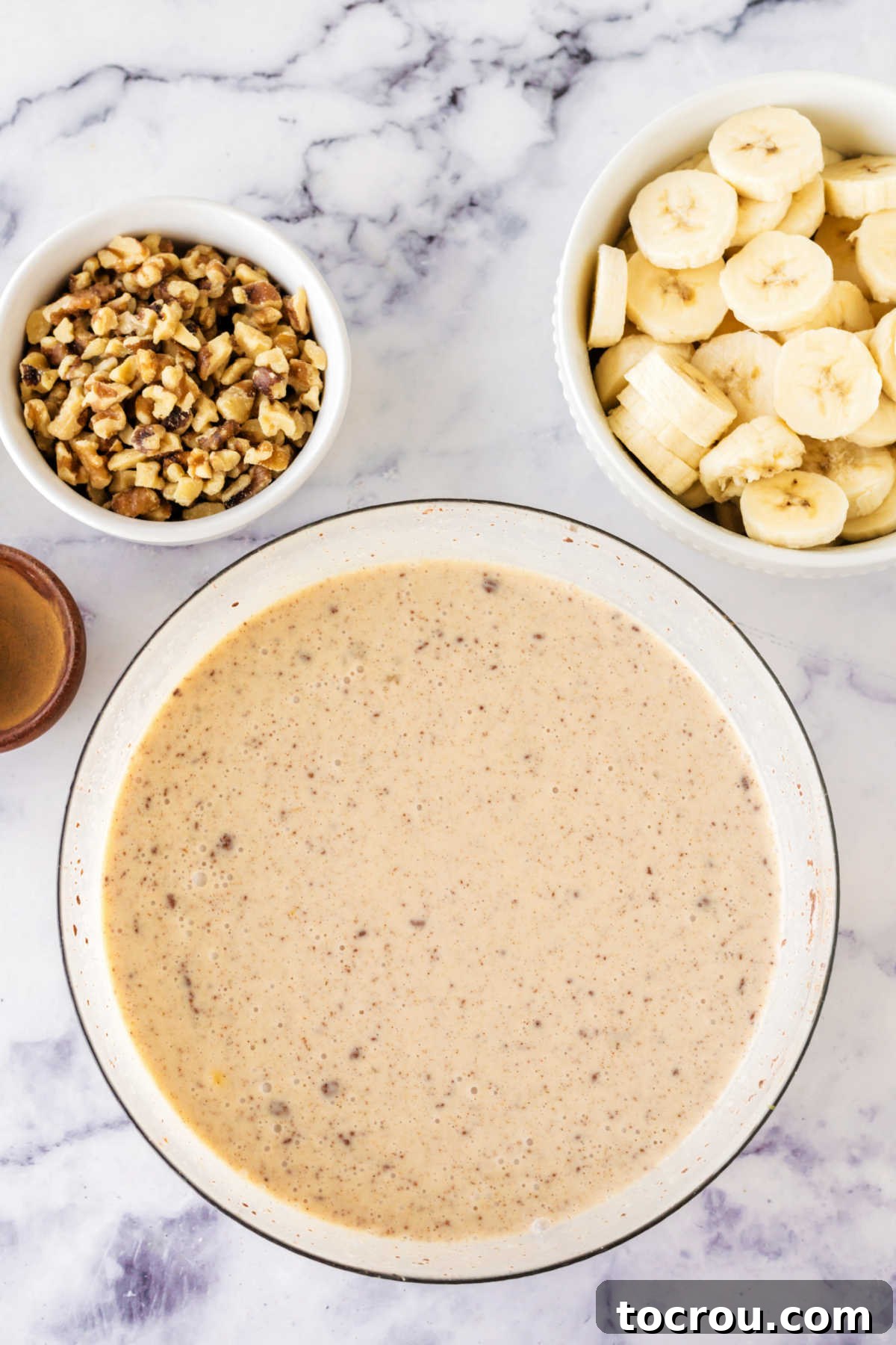 A beautifully composed shot featuring a bowl of rich cinnamon condensed milk custard positioned next to a bowl overflowing with freshly sliced bananas and another bowl of finely chopped walnuts, all prepped and ready for mixing into the bread pudding.