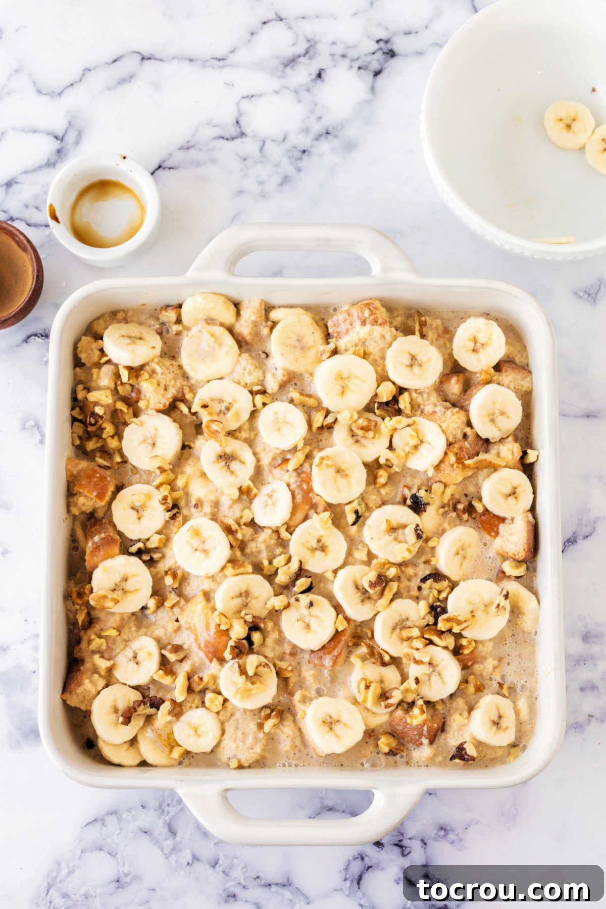A square baking dish, perfectly prepared, is brimming with a delightful mixture of bread cubes and the creamy custard, elegantly topped with artfully arranged fresh banana slices, patiently awaiting its turn to be placed in the oven.