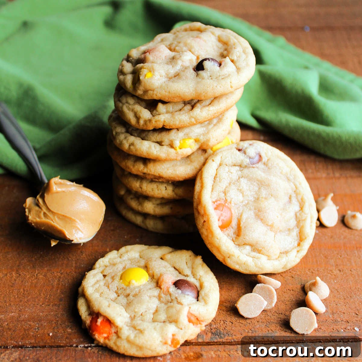 A stack of perfectly chewy triple peanut butter cookies, overflowing with peanut butter chips and Reese's Pieces. A spoonful of creamy peanut butter and scattered chips are nearby, hinting at the rich flavor.
