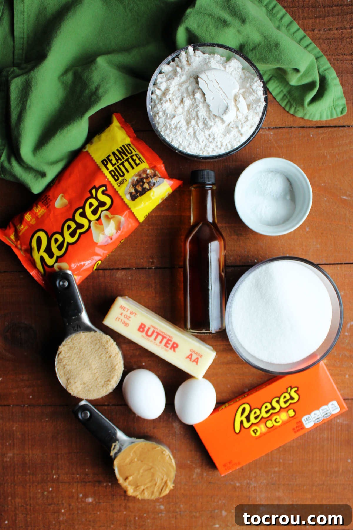 A beautiful arrangement of ingredients for triple peanut butter cookies, including a jar of peanut butter, sticks of butter, bags of flour and sugar, brown sugar, vanilla extract, fresh eggs, vibrant Reese's Pieces candies, and a bowl of peanut butter chips, all ready for baking.