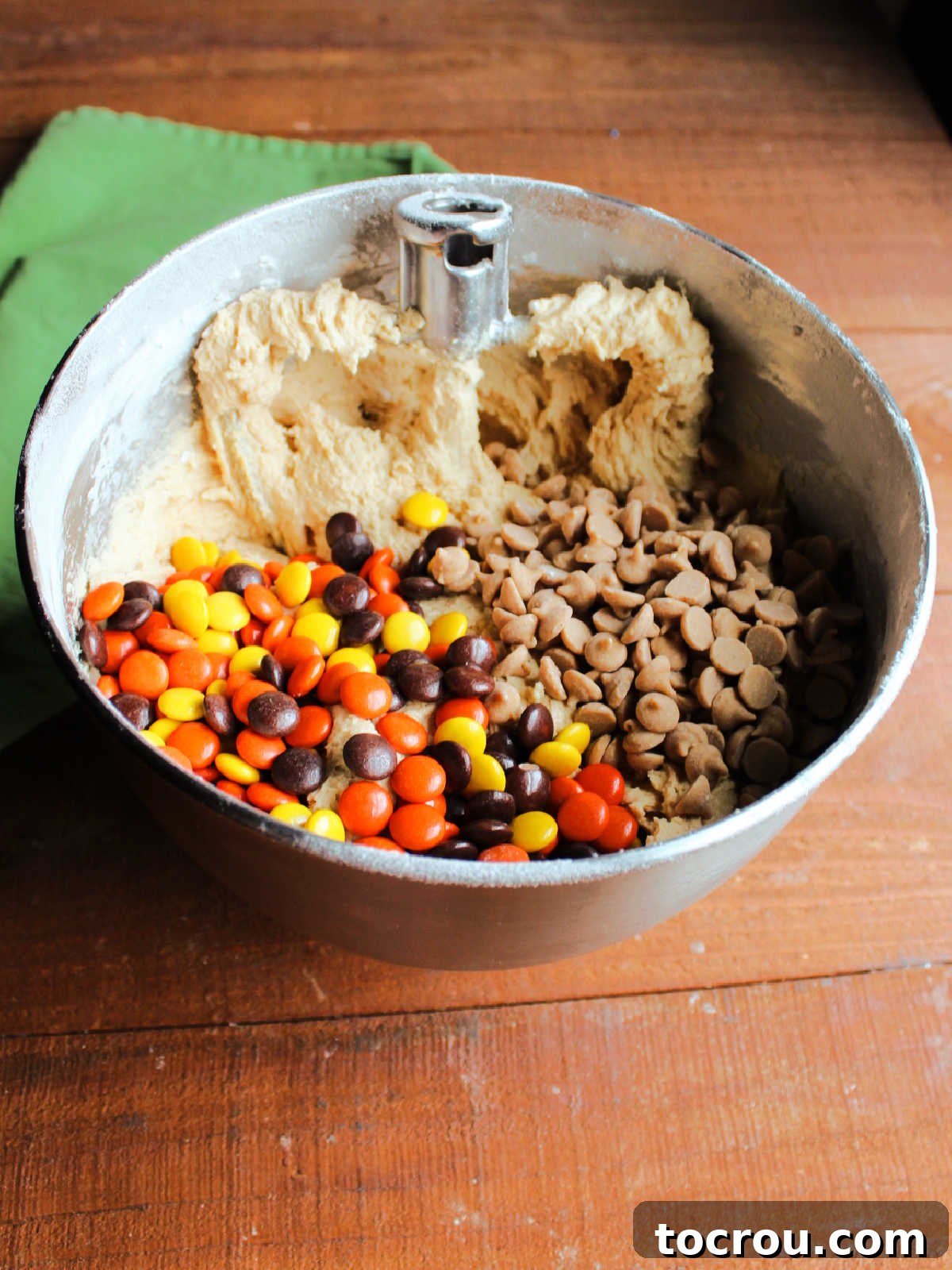 A large mixer bowl filled with soft, creamy peanut butter cookie dough, visibly laden with delicious peanut butter chips and vibrant Reese's Pieces candies, showcasing the abundant mix-ins.