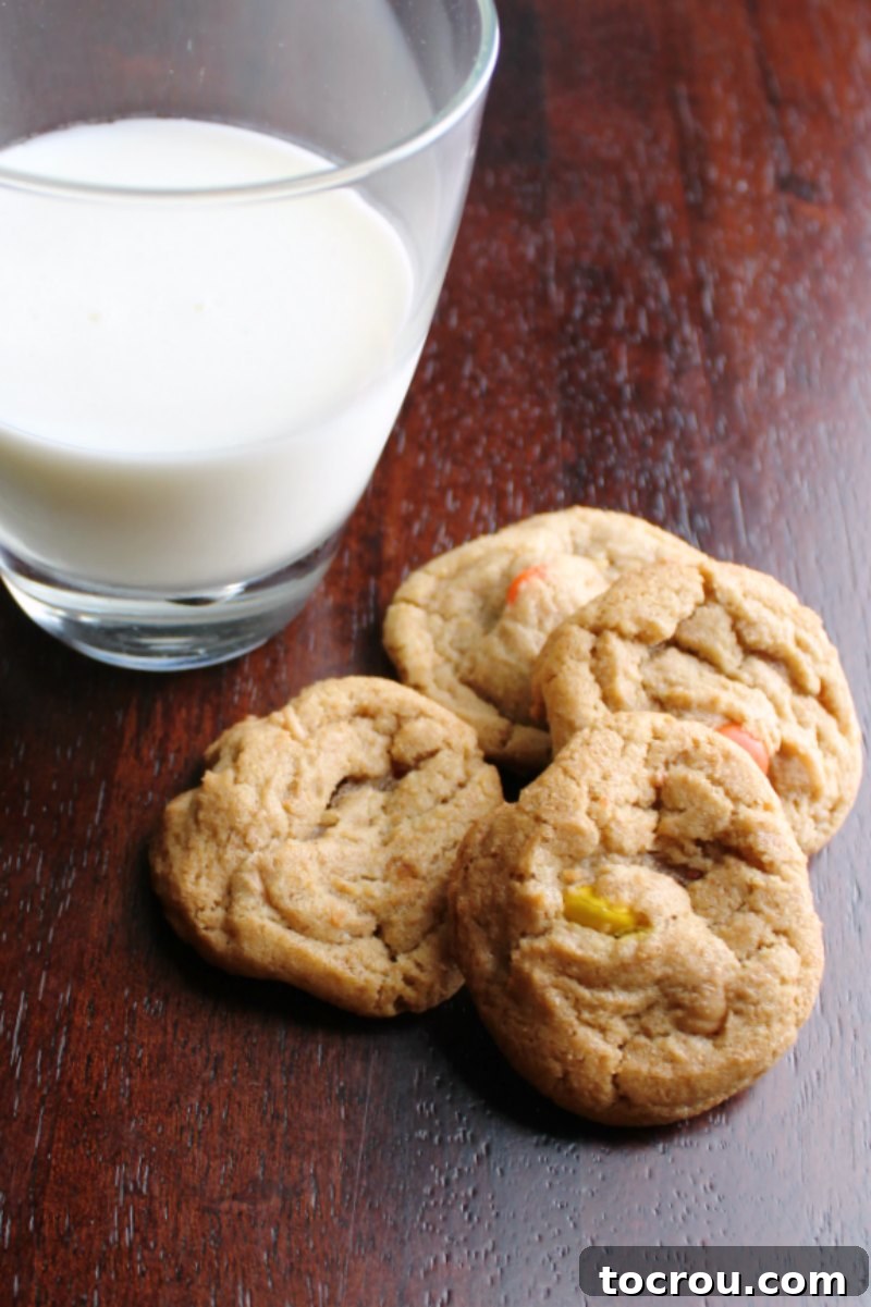 A delightful arrangement of four freshly baked triple peanut butter cookies, rich with peanut butter chips and Reese's Pieces, artfully placed next to a refreshing glass of milk, inviting a perfect pairing.