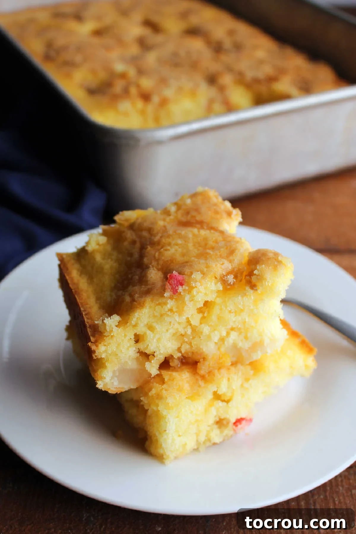 Two generous pieces of homemade fruit cocktail cake stacked enticingly on a white plate, with the remaining cake still in its baking pan visible in the soft-focus background.