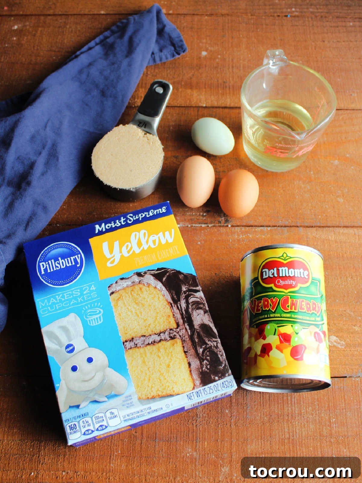 Essential ingredients for fruit cocktail cake arranged on a counter: a box of yellow cake mix, fresh eggs, a bottle of vegetable oil, a can of fruit cocktail, and a bowl of light brown sugar, all ready for baking.