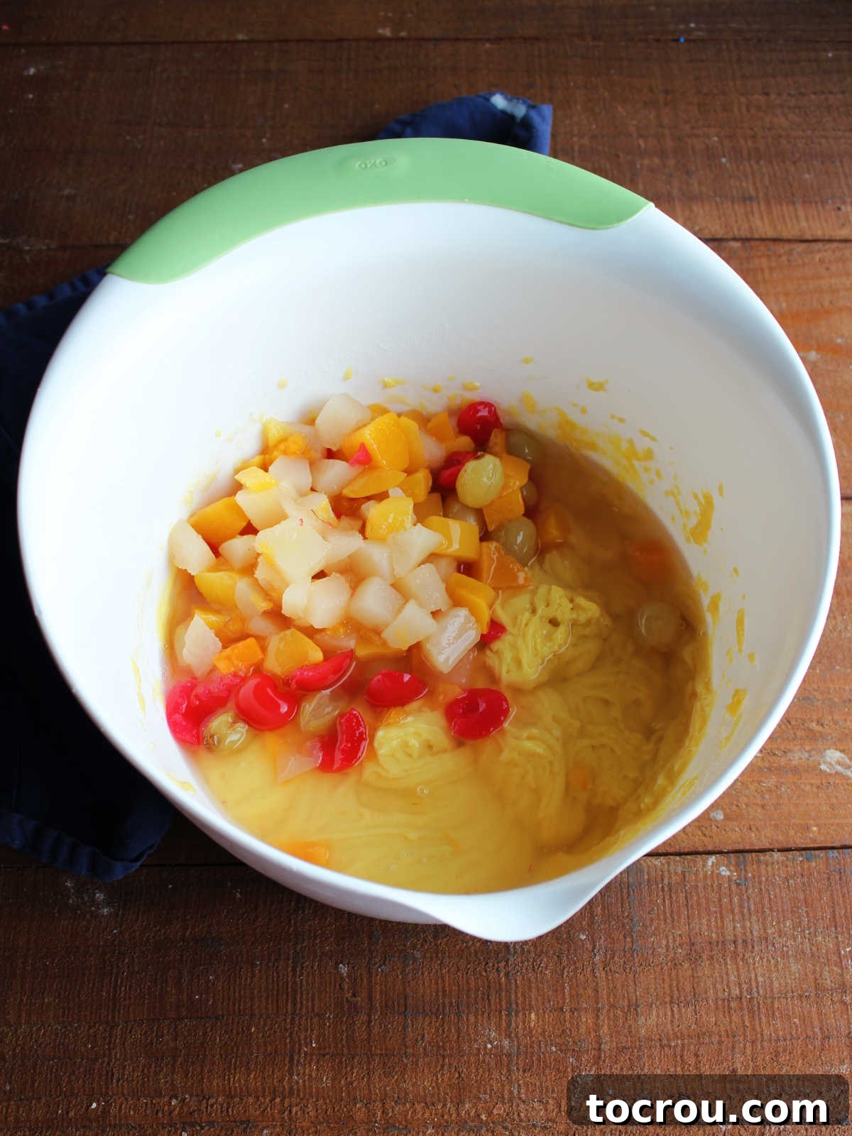 A can of colorful fruit cocktail being poured directly into a mixing bowl already containing yellow cake mix, eggs, and vegetable oil, highlighting the simple mixing process.