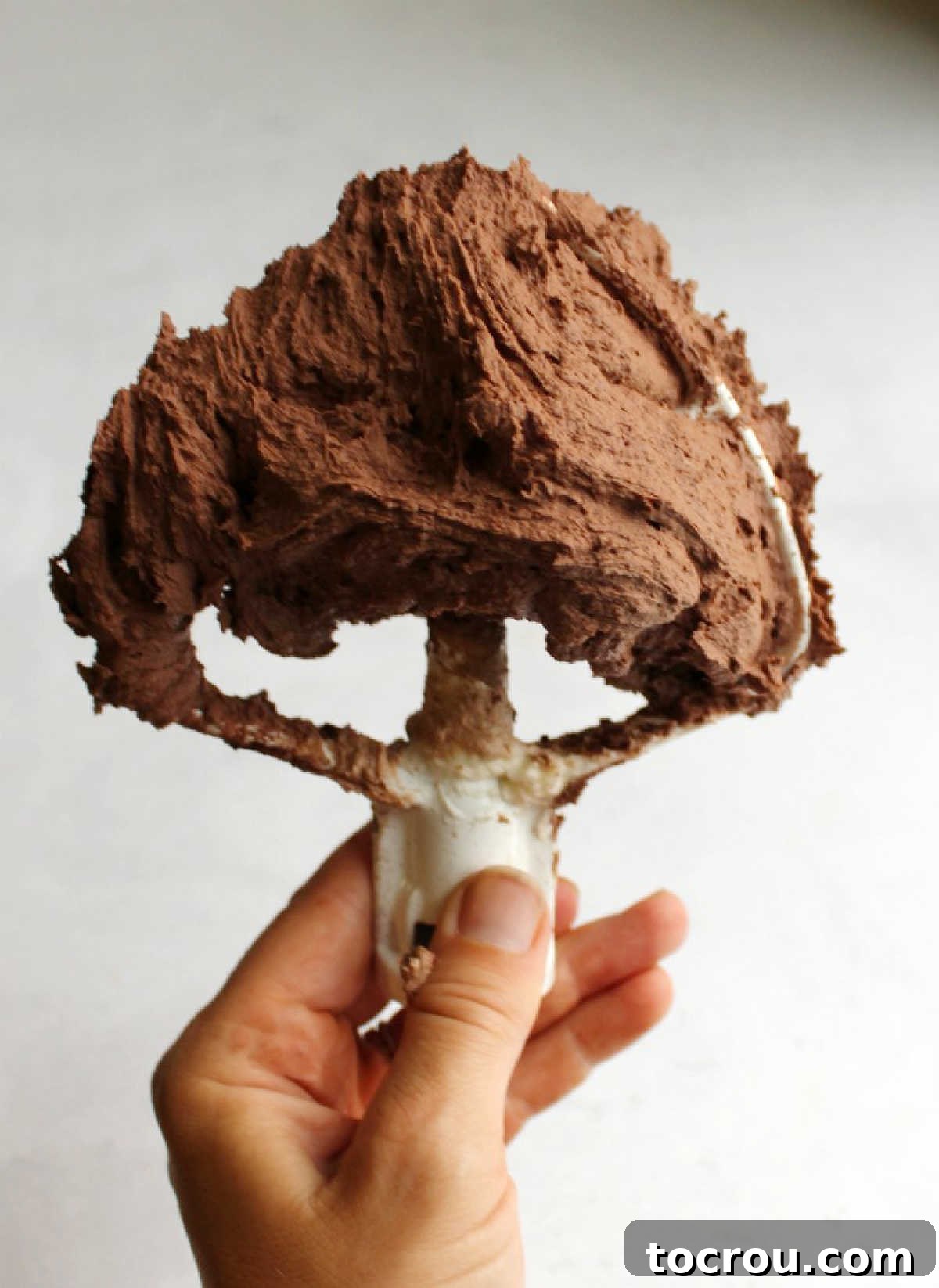 Hand holding a mixer paddle filled with dark chocolate buttercream, showcasing its rich, deep color.