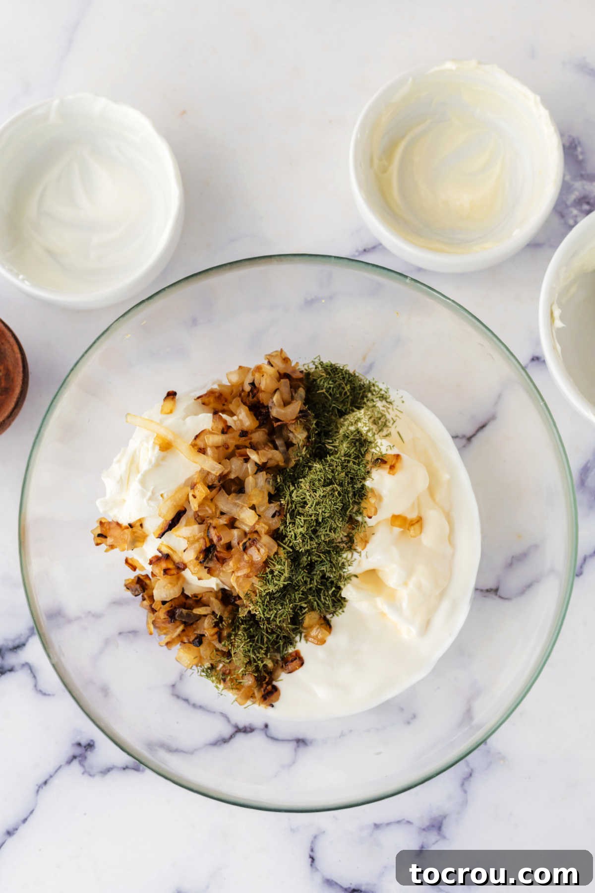 Preparing the Creamy Base with Caramelized Onions and Dill Glass mixing bowl filled with creamy mixture of sour cream, mayo, and cream cheese topped with dill and caramelized onion ready to be stirred together to make dip.