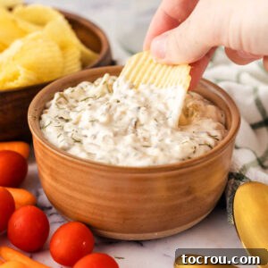Hand dipping ruffled potato chip into wood bowl filled with caramelized onion dip with baby carrots and cherry tomatoes nearby.