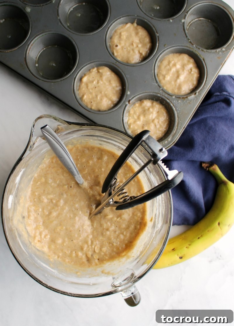 A bowl of freshly mixed banana oat muffin batter with a cookie scoop next to a muffin tin filled with batter, ready for baking.