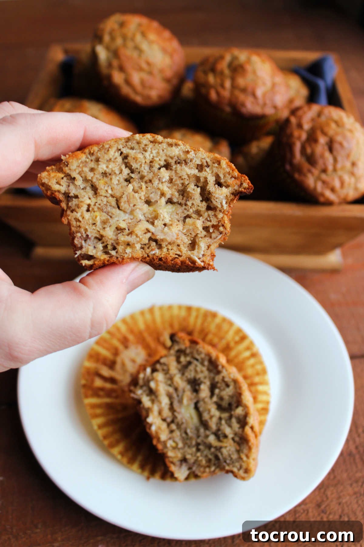 A hand gently holds half of a banana oat muffin, revealing its soft, fluffy interior filled with banana and oat textures.