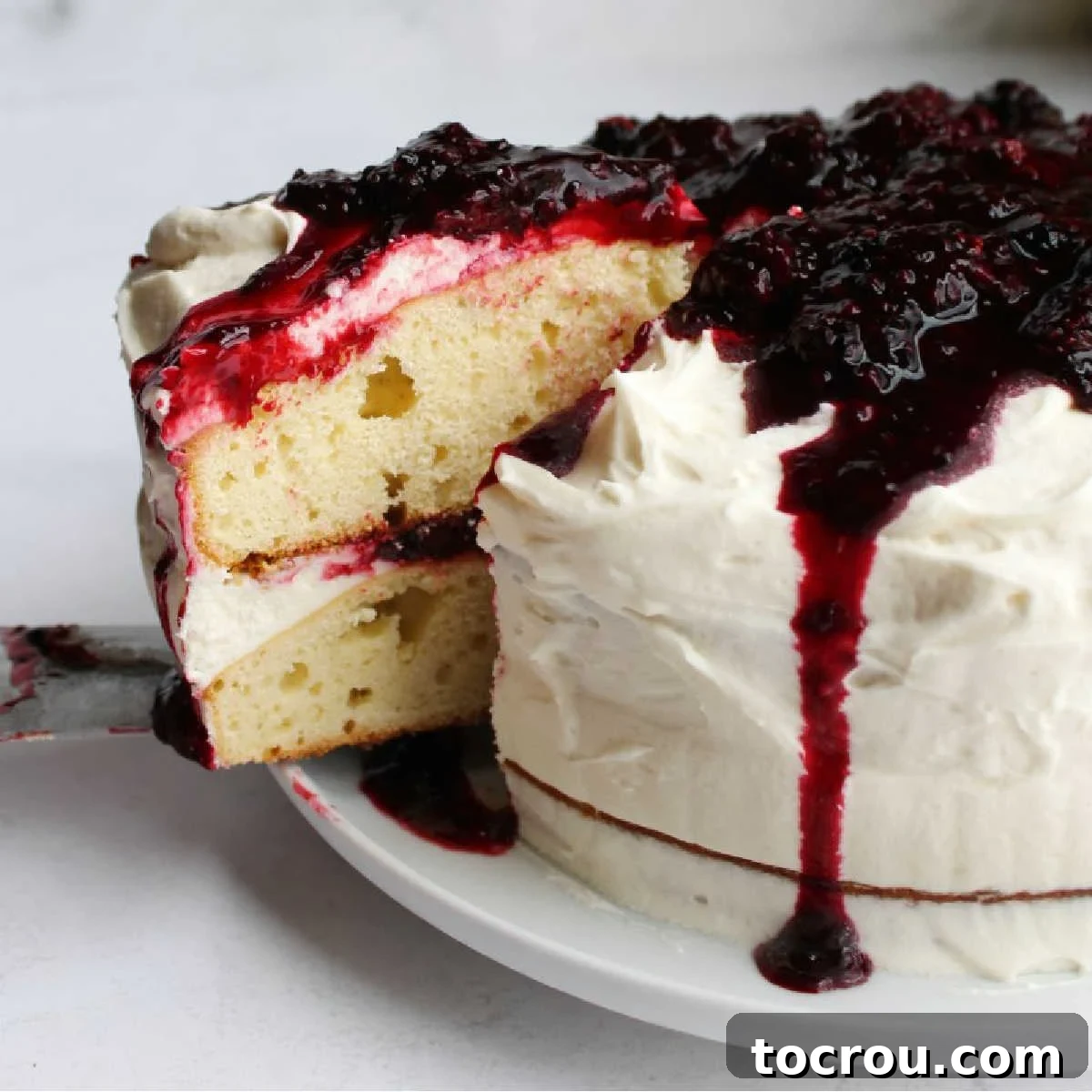 Blackberry Shortcake Bake 2 Lifting first slice of blackberry shortcake cake out showing two layers of dense cake with fluffy white ermine frosting and blackberry sauce.