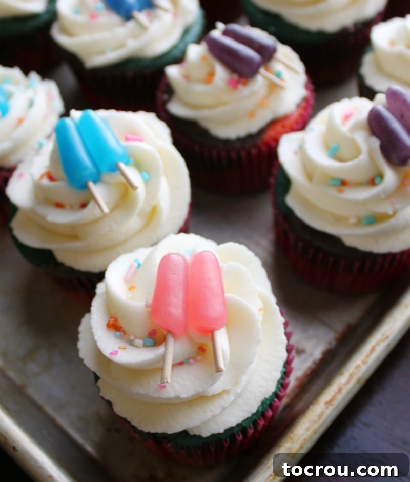 Tray of vibrant, multicolored cupcakes, generously swirled with fluffy cream cheese frosting, adorned with cheerful sprinkles, and crowned with adorable candy popsicle toppers.