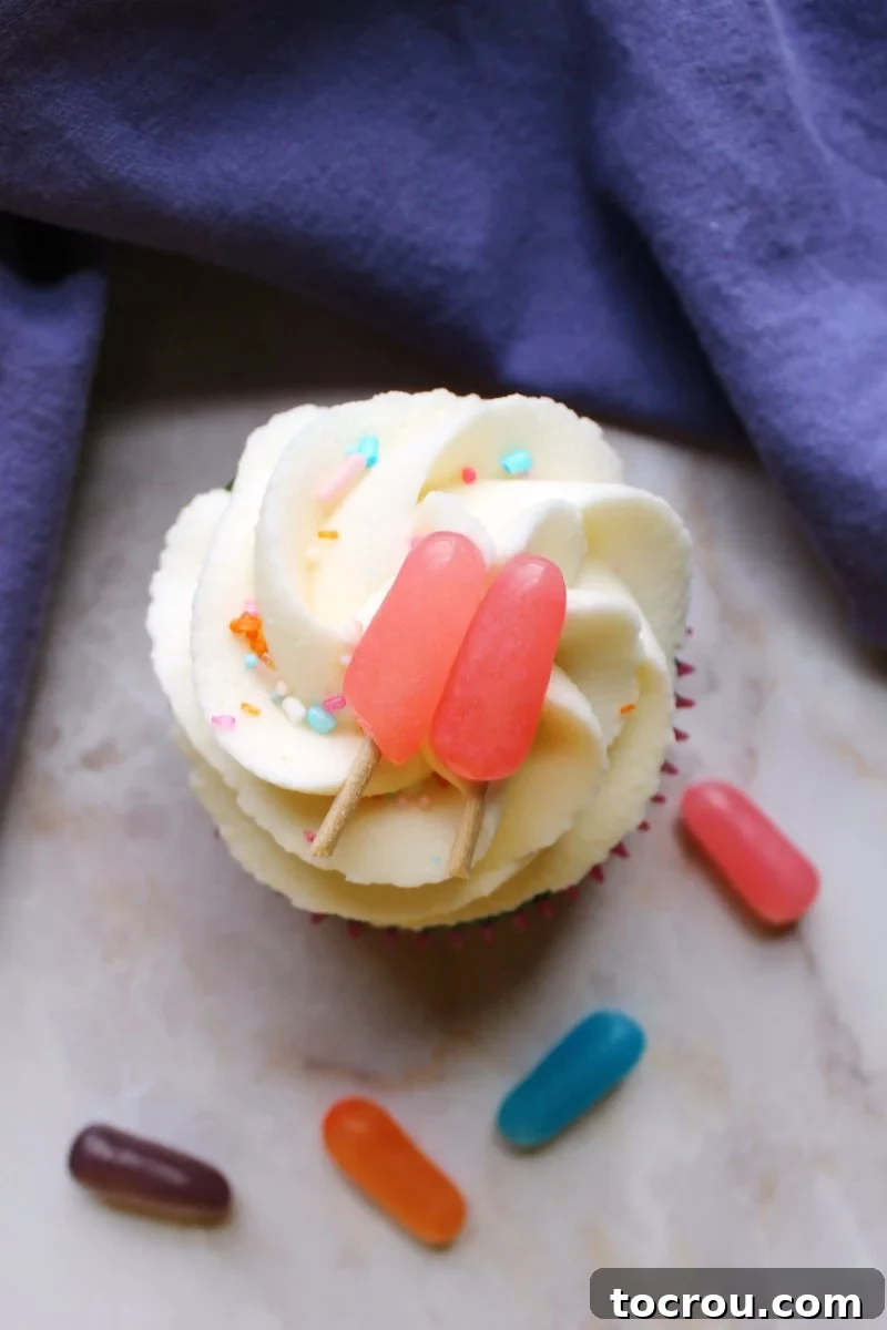 Overhead view of a single summertime cupcake featuring fluffy white frosting and a charming pink candy popsicle topper, sitting on a wooden surface.