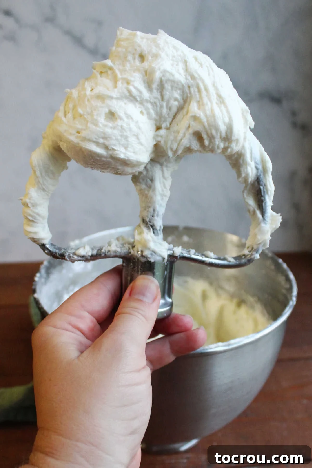 Hand holding a mixer paddle generously coated with fluffy decorator's cream cheese frosting, showcasing its smooth texture.