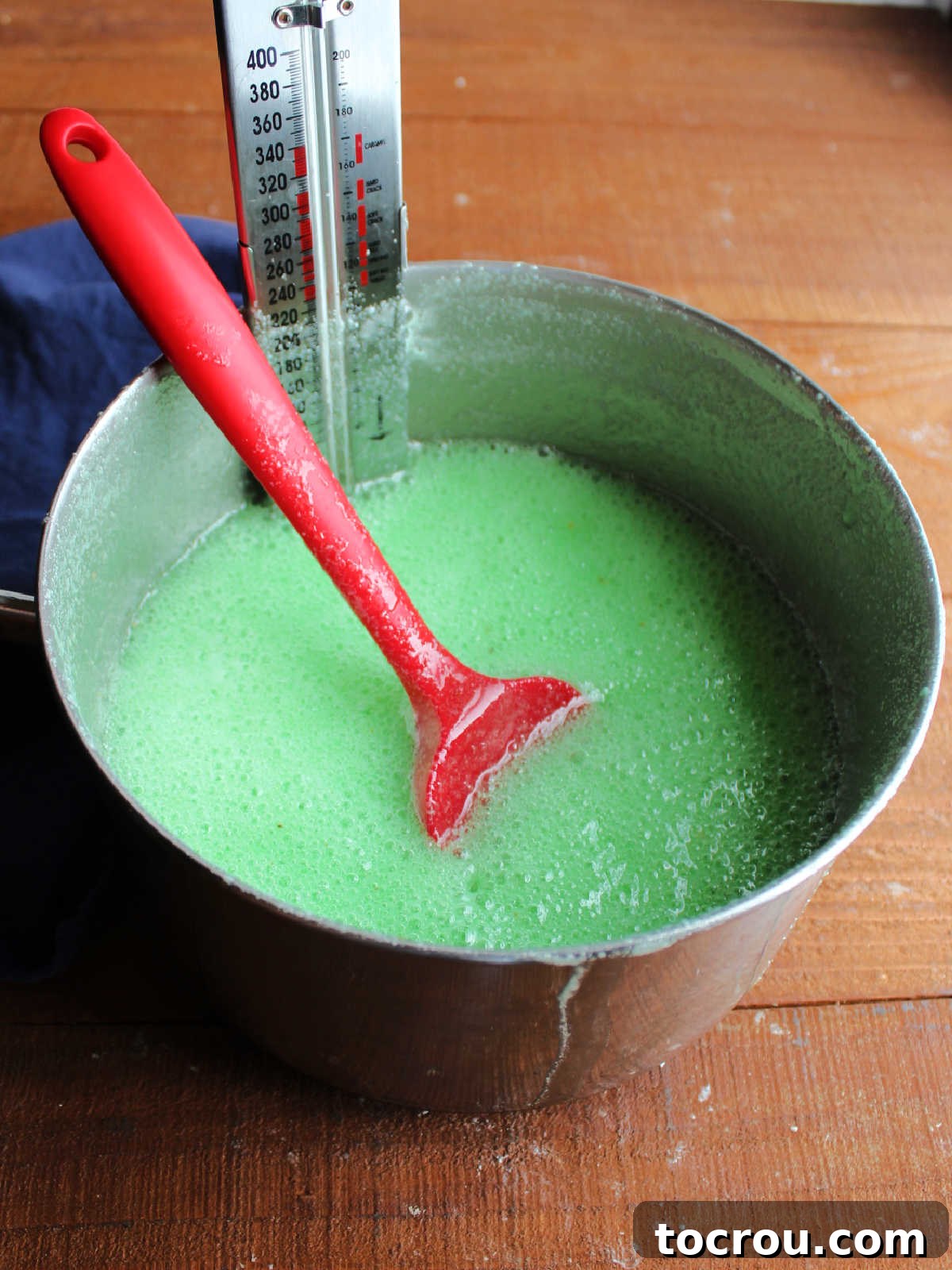 Saucepan with hot and foamy green jello mixture and candy thermometer inside.