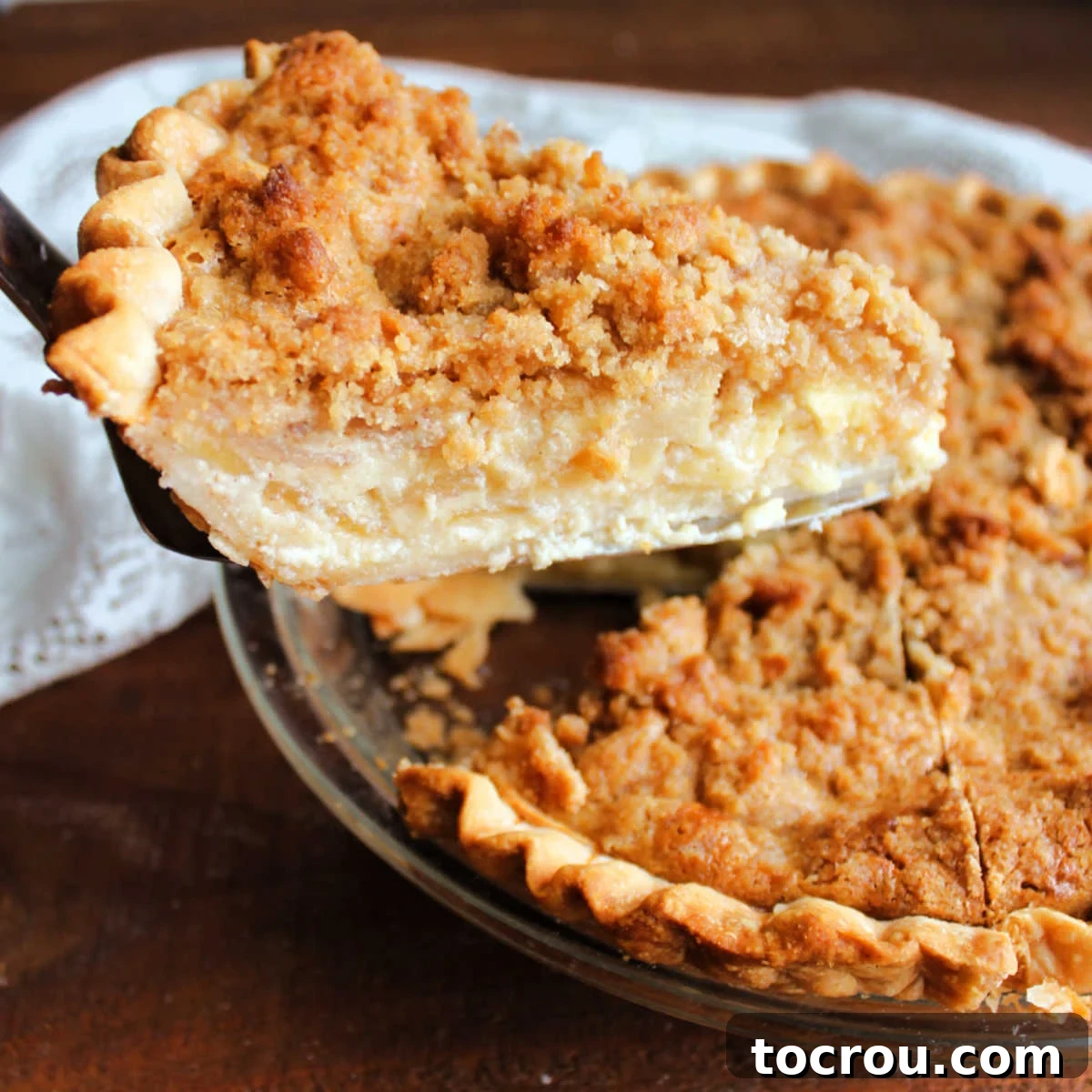 Apple Custard Crumble Pie 2 Lifting a slice of apple custard pie out of the dish showing layers of apple slices surrounded by creamy vanilla custard topped with a golden brown crumb mixture.