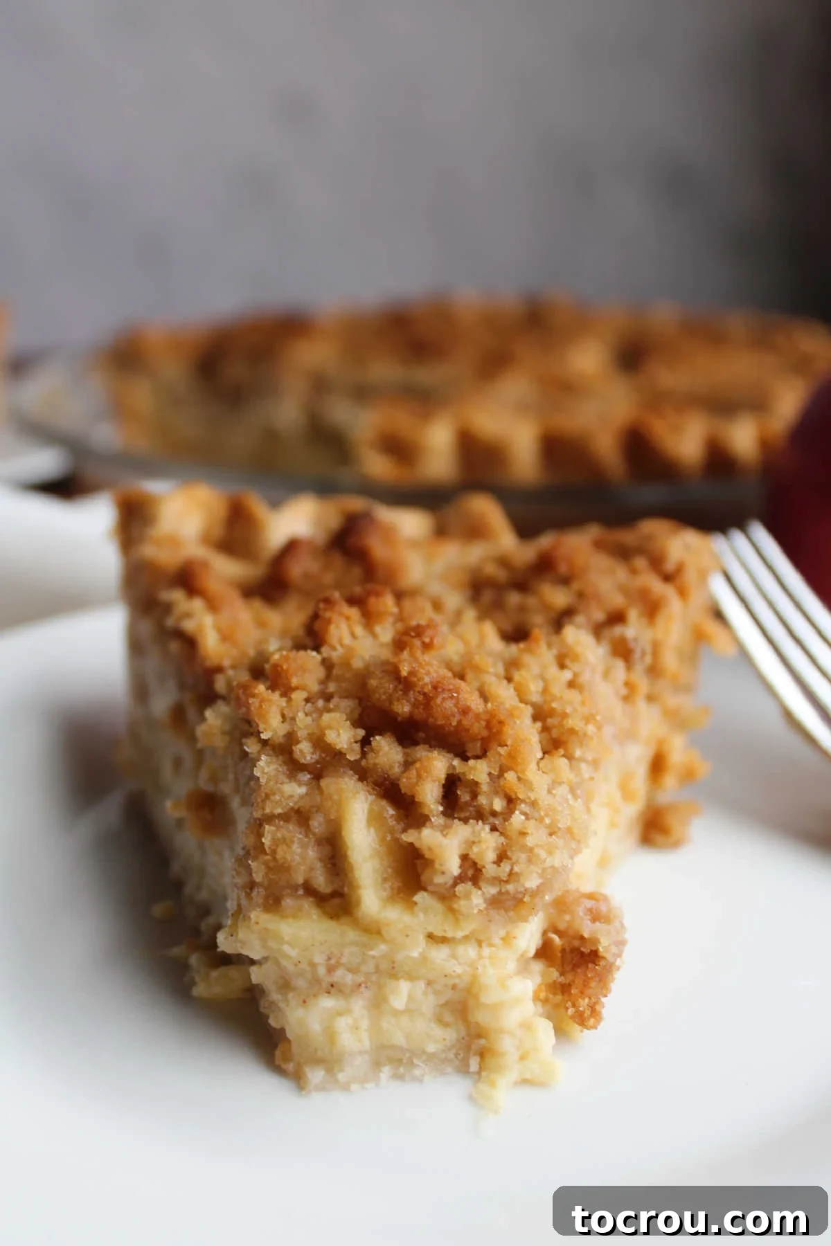 Apple Custard Crumble Pie 3 Slice of apple custard pie on plate with bite missing from tip showing creamy custard surrounding the apples and a golden streusel on top.