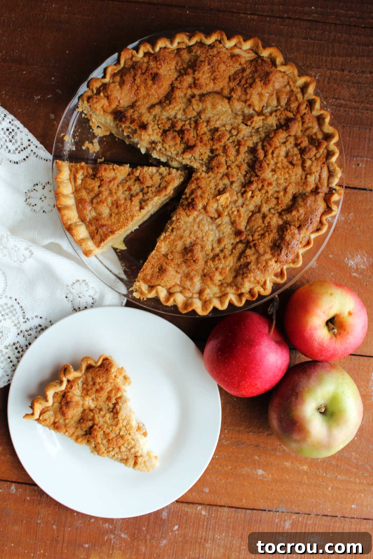 Apple Custard Crumble Pie 4 Looking down on piece of apple pie topped with golden brown butter crumb mixture next to some fresh apples and the remaining apple pie.