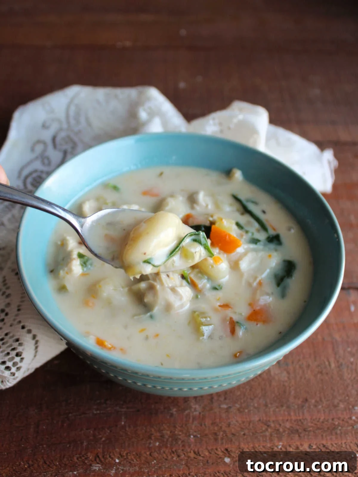Spoon being lifted out of a bowl of creamy chicken gnocchi soup with a gnocchi, some spinach and carrot on it, ready to be eaten.