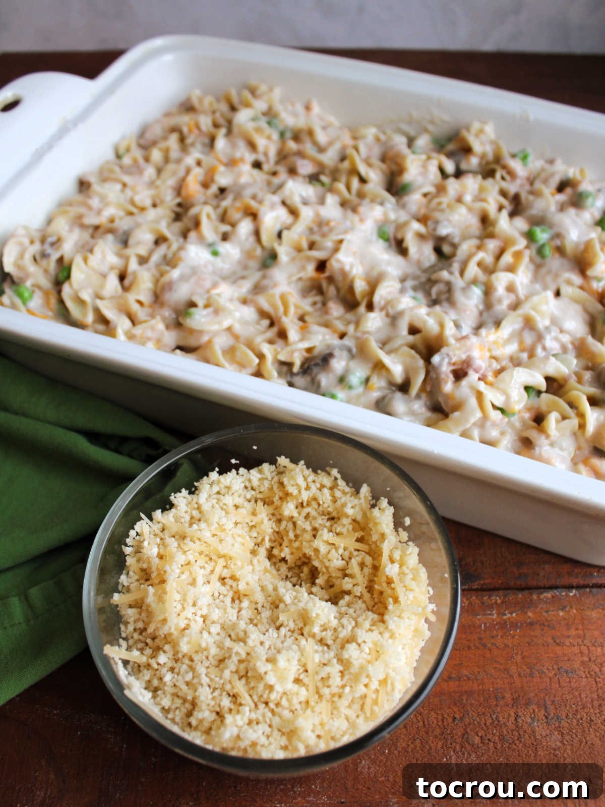 Hearty Tuna Noodle Casserole 7 Small bowl of buttered bread crumbs and Parmesan cheese sits in front of a baking pan filled with tuna noodle mixture, ready for topping.