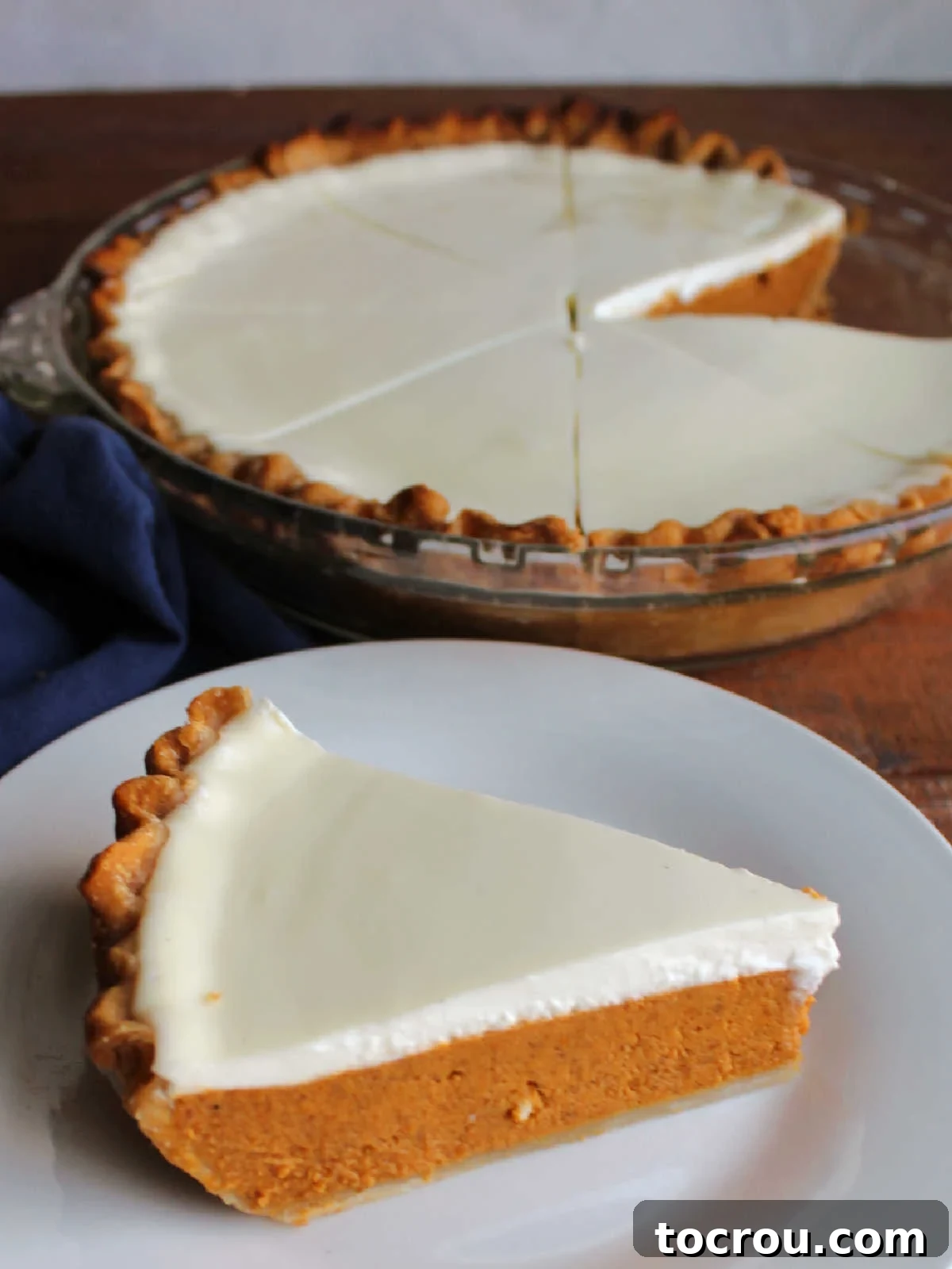 A perfect piece of layered pumpkin pie with sour cream topping served on a white plate, with the rest of the pie visible in the background.