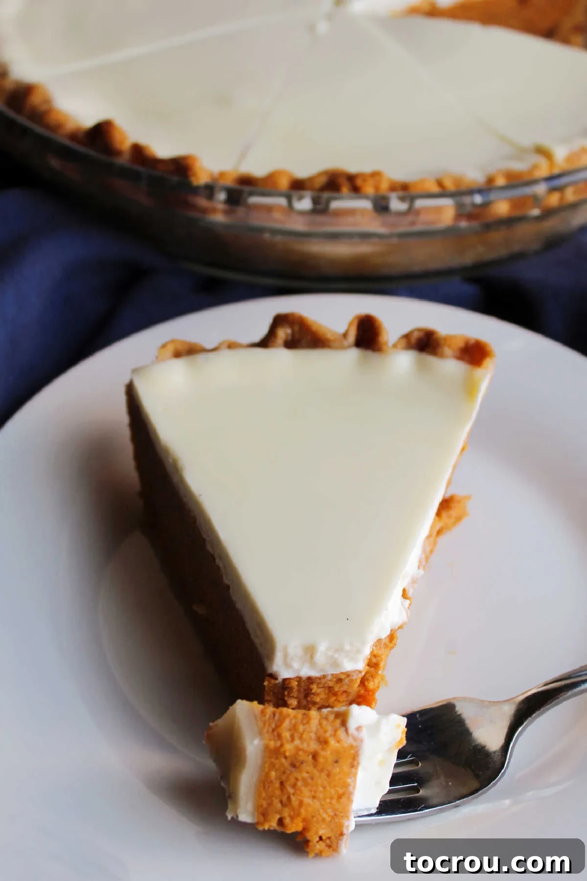A small bite of pumpkin pie on a fork, resting on a plate next to a beautifully sliced piece of pumpkin pie with its smooth sour cream topping.